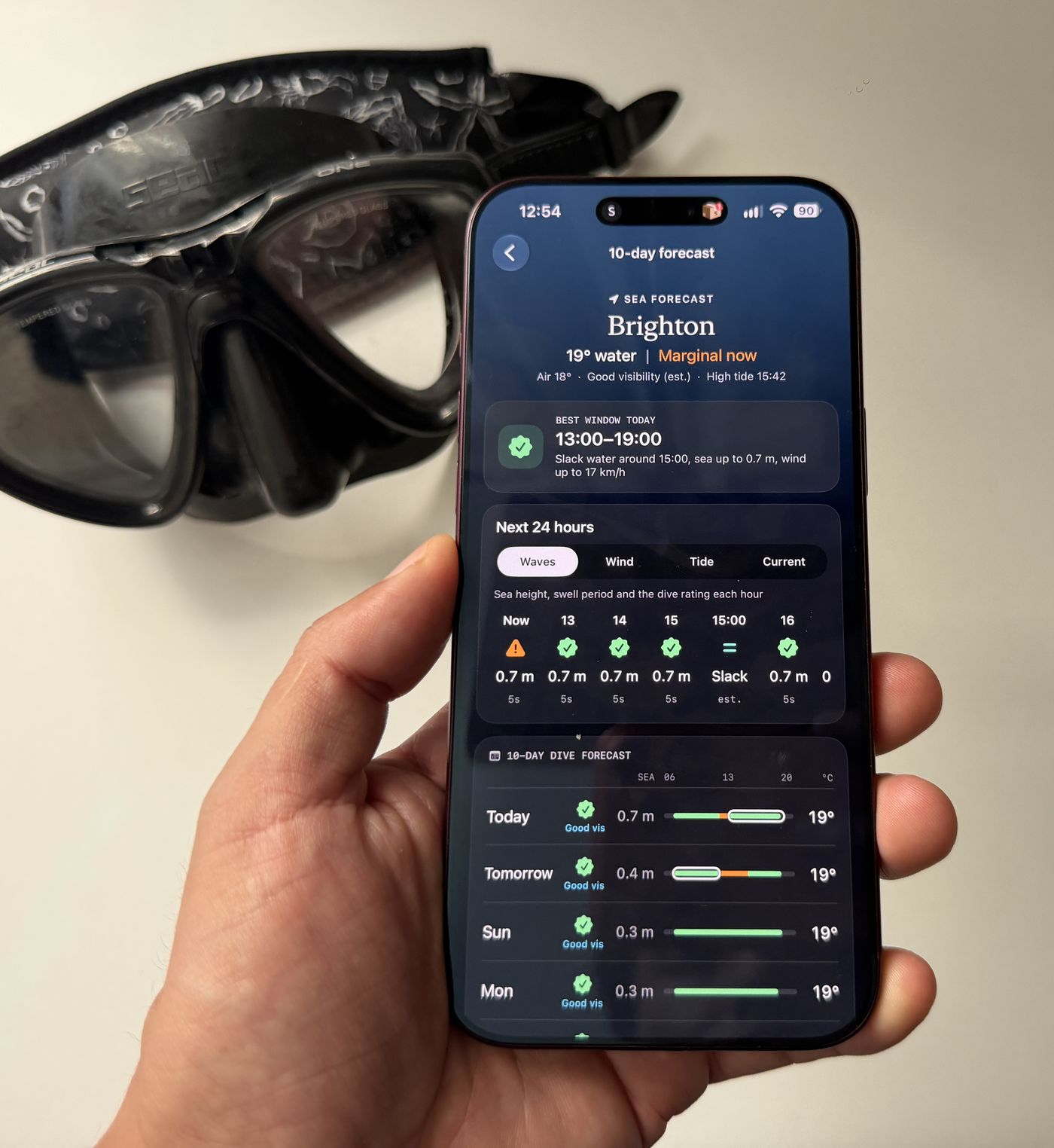 A hand holding an iPhone showing Diving Standard's 10-day sea forecast for Brighton, with the best window of the day and a rating for each day