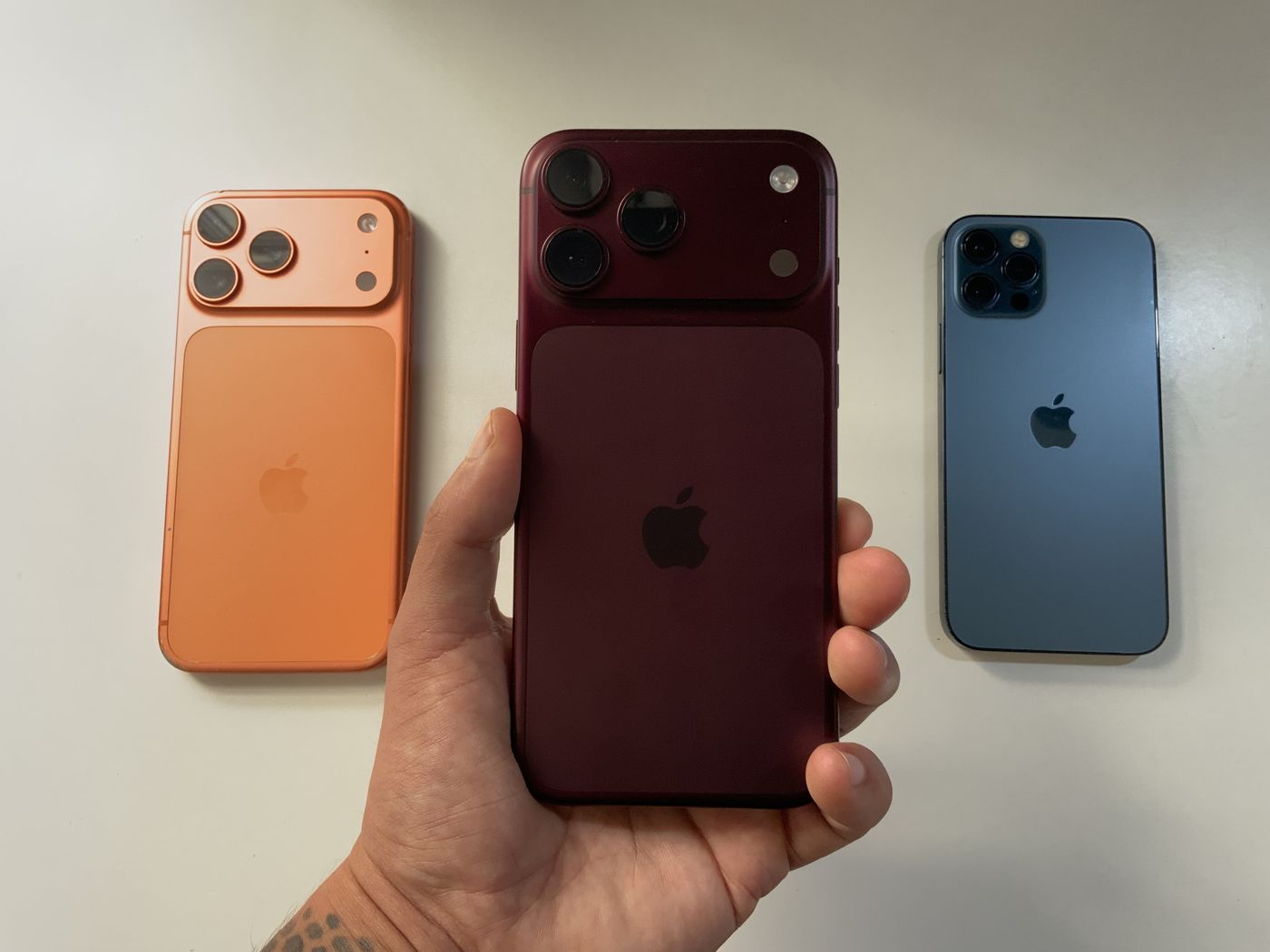 A hand holding a red iPhone 18 Pro Max in front of an orange iPhone and an older blue iPhone
