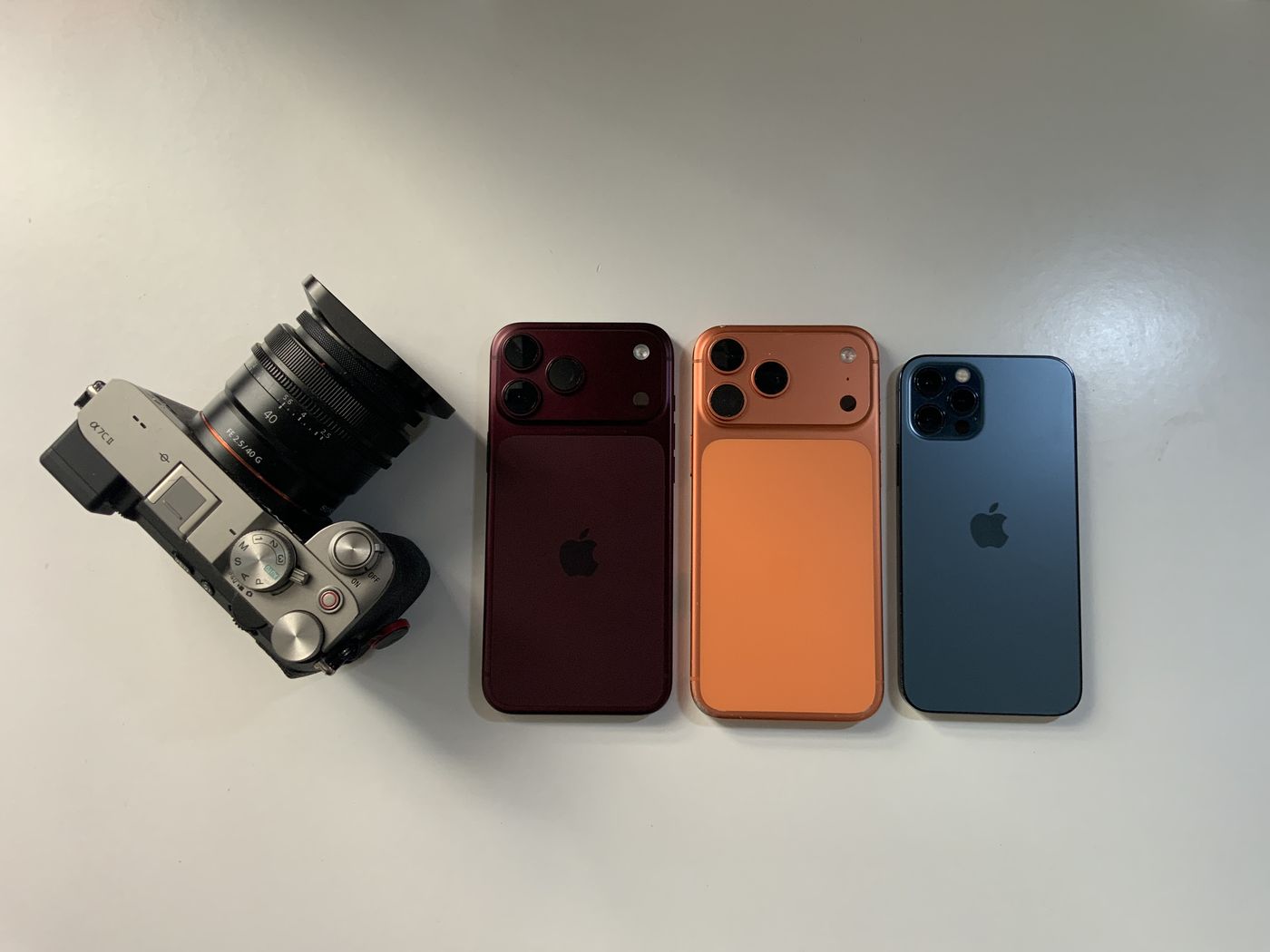 A red iPhone 18 Pro Max, two older iPhones and a mirrorless camera laid out on a white table